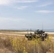 A military exercise or operation is taking place in a coastal terrain environment. An armored wheeled vehicle, consistent with a Light Armored Vehicle (LAV) or similar 8-wheeled armored personnel carrier, is positioned on or near a paved road with personnel visible around it. A single soldier in camouflage uniform is walking nearby, with the ocean visible in the background.