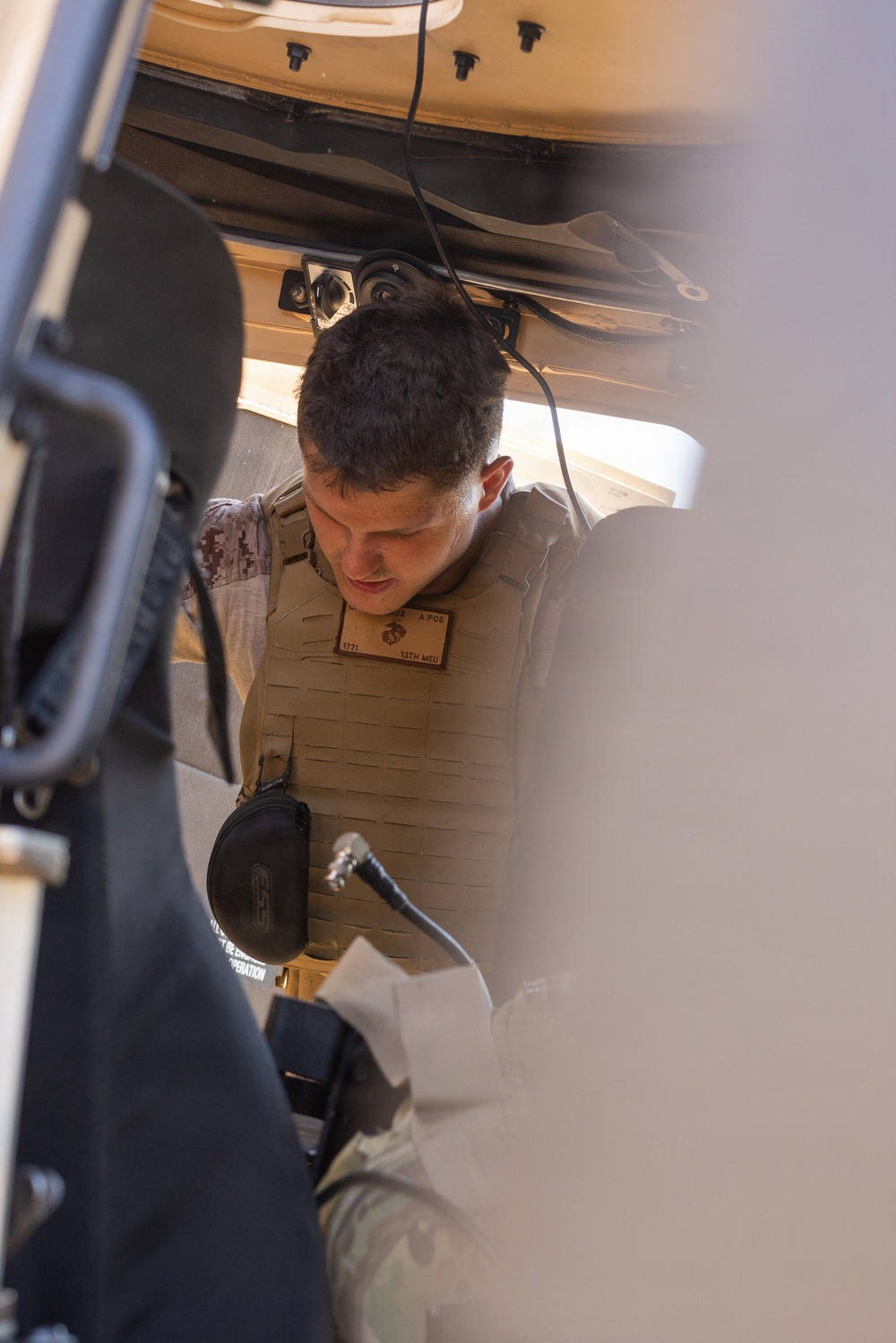 A U.S. Marine wearing a tan plate carrier vest with a USMC Eagle, Globe, and Anchor emblem and a name tape reading '13th MEU' is leaning into or inspecting a military vehicle. The individual appears to be conducting a vehicle inspection or maintenance task, viewed from inside the vehicle looking outward. The photo is taken from a low angle through a vehicle door or hatch opening.