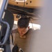 A U.S. Marine wearing a tan plate carrier vest with a USMC Eagle, Globe, and Anchor emblem and a name tape reading '13th MEU' is leaning into or inspecting a military vehicle. The individual appears to be conducting a vehicle inspection or maintenance task, viewed from inside the vehicle looking outward. The photo is taken from a low angle through a vehicle door or hatch opening.