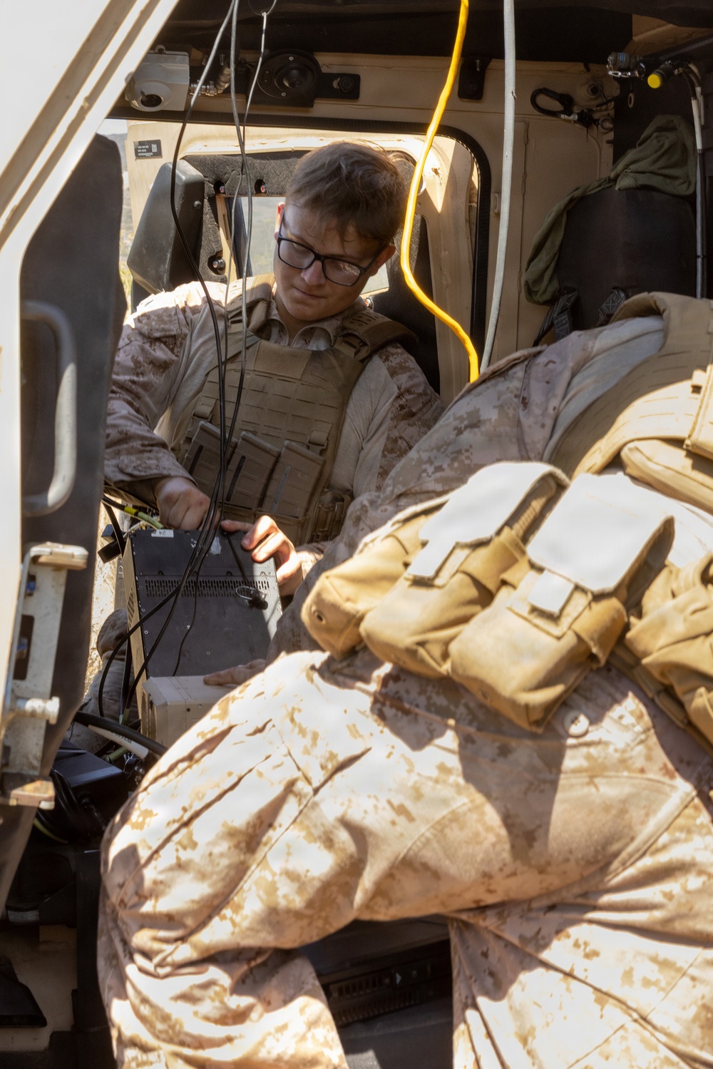 A military service member in desert MARPAT camouflage and plate carrier vest is seated inside a tactical vehicle, handling electronic equipment with multiple cables. The individual is wearing black-framed glasses and appears to be configuring or troubleshooting communications or computing hardware. A second service member in similar uniform is partially visible in the foreground.