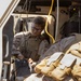 A military service member in desert MARPAT camouflage and plate carrier vest is seated inside a tactical vehicle, handling electronic equipment with multiple cables. The individual is wearing black-framed glasses and appears to be configuring or troubleshooting communications or computing hardware. A second service member in similar uniform is partially visible in the foreground.