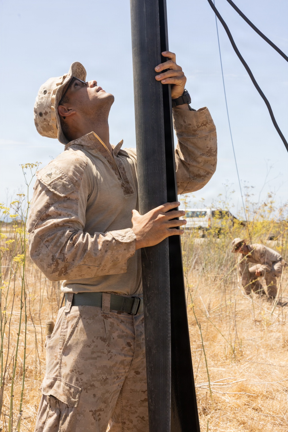 A U.S. Marine in desert MARPAT camouflage uniform is erecting or stabilizing a large black pole or mast, looking upward while gripping it with both hands. Guy-wires extend from the top of the pole, suggesting this is an antenna mast or communications/support structure being set up in a dry, arid field environment. A second service member is visible in the background, crouching and working at ground level.