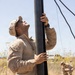 A U.S. Marine in desert MARPAT camouflage uniform is erecting or stabilizing a large black pole or mast, looking upward while gripping it with both hands. Guy-wires extend from the top of the pole, suggesting this is an antenna mast or communications/support structure being set up in a dry, arid field environment. A second service member is visible in the background, crouching and working at ground level.