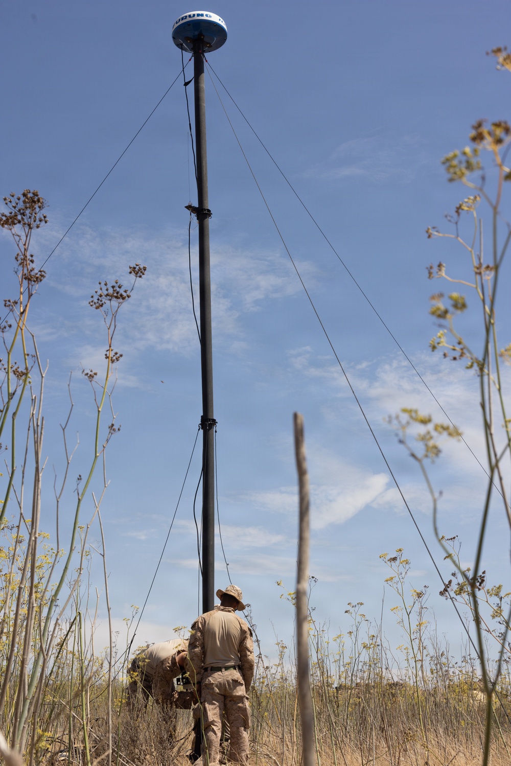 Two individuals in tan/camouflage military-style uniforms are working at the base of a tall mast or pole in an arid, scrubby outdoor environment. The mast is stabilized by multiple guy-wires and is topped with a dome-shaped antenna or sensor device. The setting appears to be a dry field with sparse, tall dried vegetation under a partly cloudy blue sky.