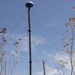 Two individuals in tan/camouflage military-style uniforms are working at the base of a tall mast or pole in an arid, scrubby outdoor environment. The mast is stabilized by multiple guy-wires and is topped with a dome-shaped antenna or sensor device. The setting appears to be a dry field with sparse, tall dried vegetation under a partly cloudy blue sky.