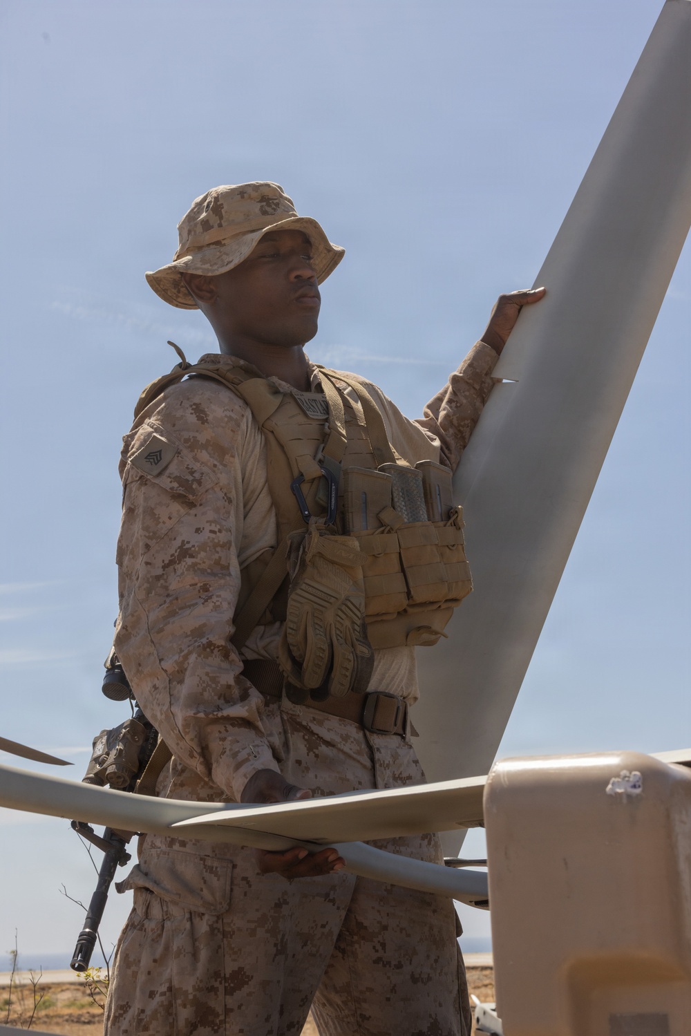 A U.S. Marine in desert MARPAT camouflage uniform and cover (boonie hat) is inspecting or handling what appears to be a rotor blade or wing component of a military aircraft or UAV in an arid, desert environment. The Marine is wearing full tactical gear including a plate carrier with magazine pouches and tactical gloves. The setting appears to be a desert training area under clear skies.