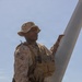 A U.S. Marine in desert MARPAT camouflage uniform and cover (boonie hat) is inspecting or handling what appears to be a rotor blade or wing component of a military aircraft or UAV in an arid, desert environment. The Marine is wearing full tactical gear including a plate carrier with magazine pouches and tactical gloves. The setting appears to be a desert training area under clear skies.