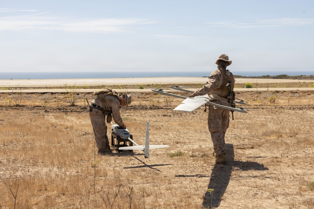 Two military personnel in desert camouflage uniforms are preparing a fixed-wing unmanned aerial vehicle (UAV) for launch in an arid field near a coastal area. One soldier holds the UAV airframe while the other tends to ground equipment, suggesting a pre-launch setup operation. The background reveals a road, sparse dry vegetation, and a visible coastline or large body of water.