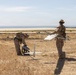 Two military personnel in desert camouflage uniforms are preparing a fixed-wing unmanned aerial vehicle (UAV) for launch in an arid field near a coastal area. One soldier holds the UAV airframe while the other tends to ground equipment, suggesting a pre-launch setup operation. The background reveals a road, sparse dry vegetation, and a visible coastline or large body of water.