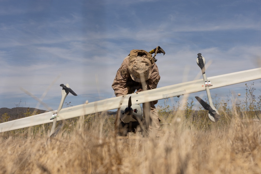A military service member in desert camouflage (MARPAT pattern) is crouched over a fixed-wing unmanned aerial vehicle (UAV) in an open field of dry grass. The individual appears to be preparing, assembling, or inspecting the drone prior to or following a flight operation. The setting appears to be an arid training environment with hills visible in the background.