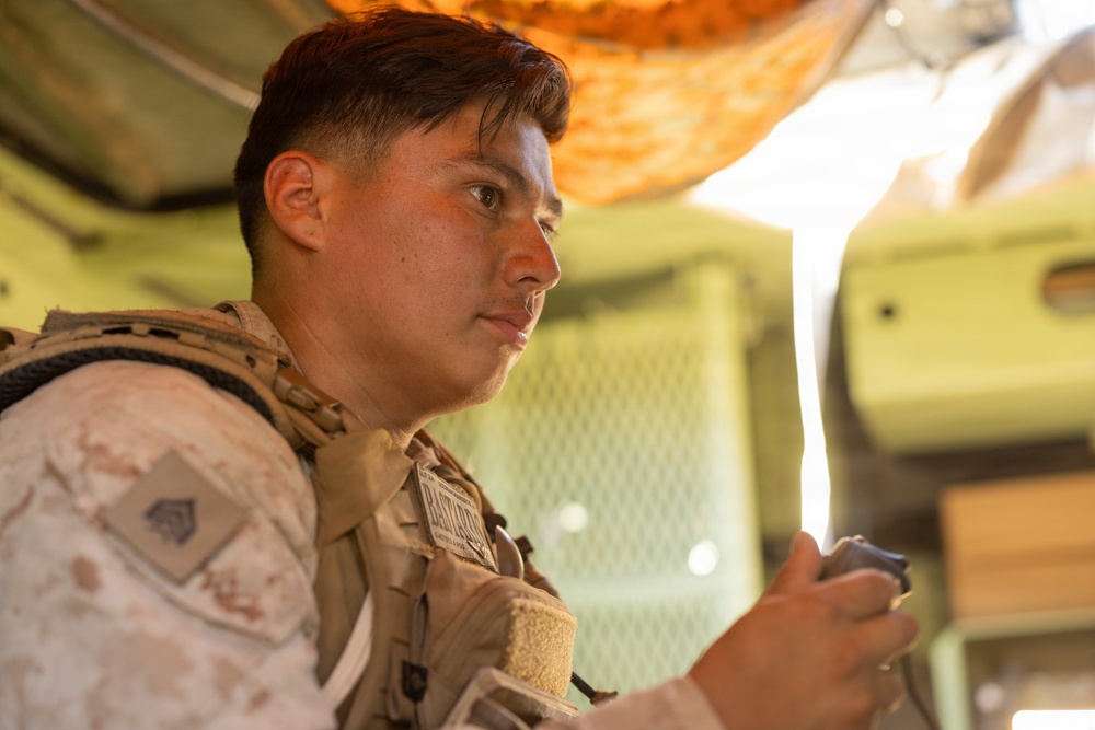 A young male service member in U.S. Marine Corps desert MARPAT camouflage uniform and tactical vest is photographed in a three-quarter profile view. He appears to be inside a military vehicle or structure, holding a small device in his hand. The image is a close-up portrait with shallow depth of field and warm lighting.