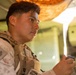 A young male service member in U.S. Marine Corps desert MARPAT camouflage uniform and tactical vest is photographed in a three-quarter profile view. He appears to be inside a military vehicle or structure, holding a small device in his hand. The image is a close-up portrait with shallow depth of field and warm lighting.