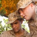 Two military personnel wearing desert camouflage uniforms (MARPAT desert pattern) are shown in close proximity, apparently reviewing something together beneath tree cover. Both wear covers (caps) with what appears to be the Eagle, Globe, and Anchor emblem visible on one cap, indicating U.S. Marine Corps affiliation. The setting appears to be an outdoor field environment with foliage providing overhead concealment.