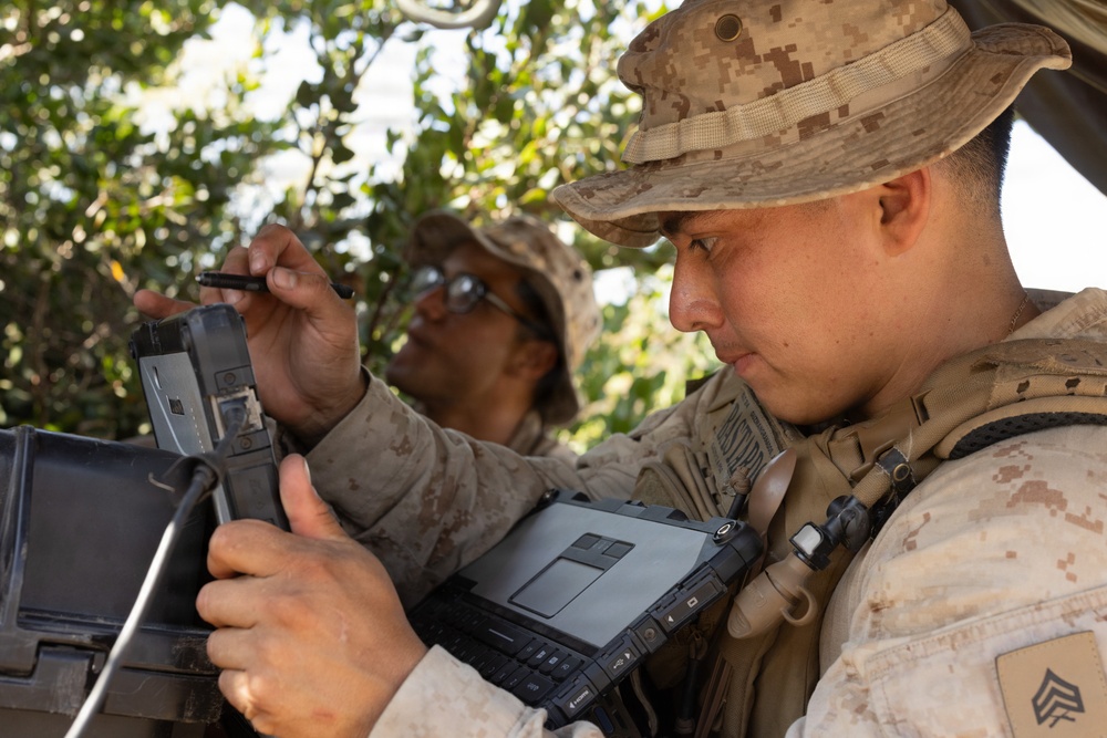 Two U.S. Marines in desert MARPAT camouflage uniforms are operating ruggedized military tablet/computing devices in a field environment. The foreground Marine, identifiable by sergeant chevrons on his collar, is holding a rugged laptop/tablet while the second Marine uses a stylus on a separate device. They appear to be conducting a field exercise or operational task involving digital equipment.