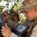 Two U.S. Marines in desert MARPAT camouflage uniforms are operating ruggedized military tablet/computing devices in a field environment. The foreground Marine, identifiable by sergeant chevrons on his collar, is holding a rugged laptop/tablet while the second Marine uses a stylus on a separate device. They appear to be conducting a field exercise or operational task involving digital equipment.