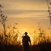 A silhouetted figure walks away from the camera along a narrow dirt path through dry grassland and tall weeds during golden hour. The warm orange and yellow sunset sky creates a dramatic backlit effect, rendering the person and surrounding vegetation as dark silhouettes. The figure appears to be carrying equipment, possibly a backpack and a long implement such as a rifle or walking staff.