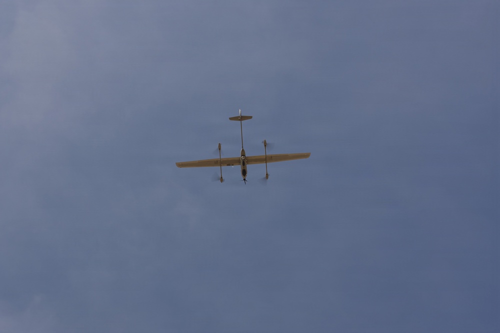 The image shows a small fixed-wing unmanned aerial vehicle (UAV) or drone photographed from below against an overcast blue-grey sky. The aircraft appears to be in level flight and is viewed from a ground-level perspective looking upward. The aircraft has a distinctive twin-boom or pusher-propeller configuration visible from the underside.
