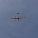 The image shows a small fixed-wing unmanned aerial vehicle (UAV) or drone photographed from below against an overcast blue-grey sky. The aircraft appears to be in level flight and is viewed from a ground-level perspective looking upward. The aircraft has a distinctive twin-boom or pusher-propeller configuration visible from the underside.