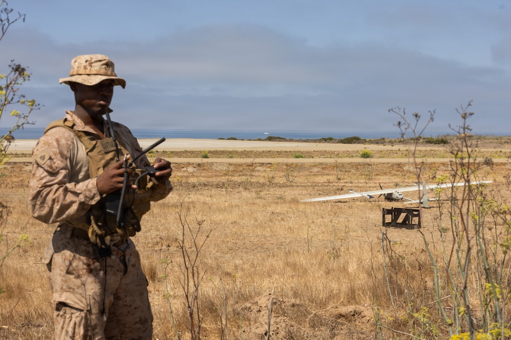 A military service member in desert camouflage (MARPAT) uniform operates a remote control device in an open field near a coastline. In the background, a fixed-wing unmanned aerial vehicle (UAV) is visible on what appears to be a launch rail or ground support equipment. The setting appears to be a coastal training area with dry grassland vegetation.