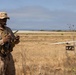 A military service member in desert camouflage (MARPAT) uniform operates a remote control device in an open field near a coastline. In the background, a fixed-wing unmanned aerial vehicle (UAV) is visible on what appears to be a launch rail or ground support equipment. The setting appears to be a coastal training area with dry grassland vegetation.