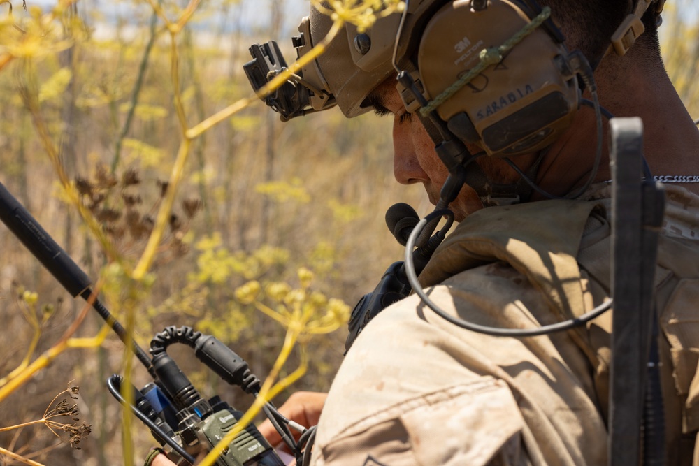 A military service member wearing tactical gear is operating a radio communication device in a field environment. The individual is equipped with a combat helmet featuring night vision goggle mounts, tactical hearing protection headset with boom microphone, and is actively using a handheld radio. The background consists of dry, yellow-brown vegetation consistent with an arid or late-season outdoor training or operational environment.