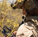 A military service member wearing tactical gear is operating a radio communication device in a field environment. The individual is equipped with a combat helmet featuring night vision goggle mounts, tactical hearing protection headset with boom microphone, and is actively using a handheld radio. The background consists of dry, yellow-brown vegetation consistent with an arid or late-season outdoor training or operational environment.