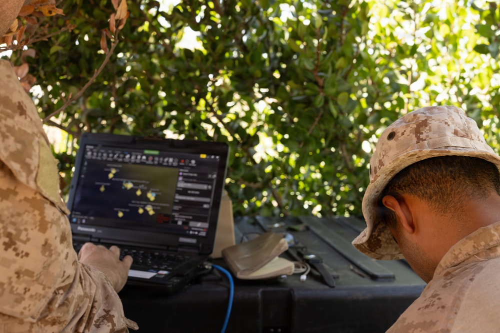 Two military personnel in desert camouflage uniforms are operating a ruggedized Panasonic laptop displaying what appears to be a tactical mapping or command-and-control software interface. They are positioned outdoors under tree cover, working at a field table with additional equipment visible. The setting suggests a field operations or command post environment.
