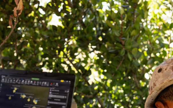 Two military personnel in desert camouflage uniforms are operating a ruggedized Panasonic laptop displaying what appears to be a tactical mapping or command-and-control software interface. They are positioned outdoors under tree cover, working at a field table with additional equipment visible. The setting suggests a field operations or command post environment.