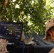 Two military personnel in desert camouflage uniforms are operating a ruggedized Panasonic laptop displaying what appears to be a tactical mapping or command-and-control software interface. They are positioned outdoors under tree cover, working at a field table with additional equipment visible. The setting suggests a field operations or command post environment.