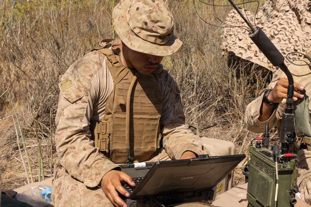 A military service member in desert camouflage (MARPAT pattern) is operating a ruggedized Getac laptop in an outdoor field environment. A second service member is visible in the background handling a radio antenna mounted on communications equipment. The scene appears to depict a field communications or command post setup during a training exercise or operational deployment.