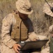 A military service member in desert camouflage (MARPAT pattern) is operating a ruggedized Getac laptop in an outdoor field environment. A second service member is visible in the background handling a radio antenna mounted on communications equipment. The scene appears to depict a field communications or command post setup during a training exercise or operational deployment.