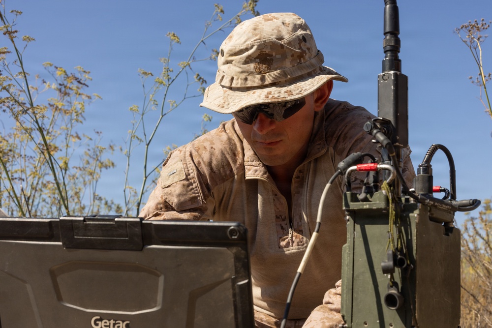A military service member in desert camouflage (MARPAT pattern) is operating a ruggedized Getac laptop connected to what appears to be a military radio or communications device in an outdoor field environment. The individual is wearing a boonie hat and sunglasses, suggesting operations in a sunny, arid climate. The setup indicates a field communications or command-and-control operation.