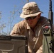 A military service member in desert camouflage (MARPAT pattern) is operating a ruggedized Getac laptop connected to what appears to be a military radio or communications device in an outdoor field environment. The individual is wearing a boonie hat and sunglasses, suggesting operations in a sunny, arid climate. The setup indicates a field communications or command-and-control operation.