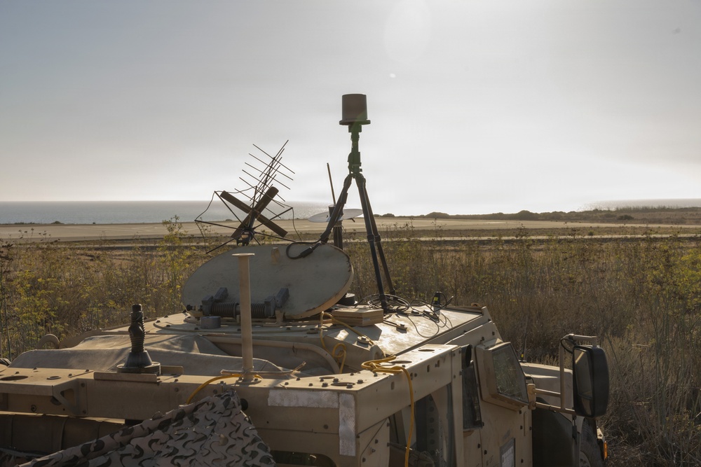 A military tactical vehicle, likely a HMMWV (Humvee), is equipped with multiple communications and surveillance systems mounted on its roof. The vehicle is positioned in an open coastal terrain environment, partially concealed with camouflage netting. The setup suggests a forward command, communications, or electronic warfare post.