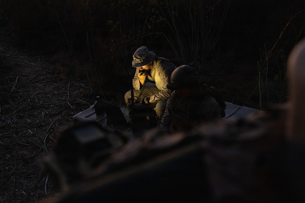 A nighttime military field operation showing at least two personnel in camouflage uniforms gathered around equipment in a wooded area. One individual wearing a boonie hat is illuminated by a light source, appearing to use a radio handset or communication device. The scene suggests a tactical communications or command post activity conducted under low-light conditions.