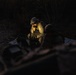 A nighttime military field operation showing at least two personnel in camouflage uniforms gathered around equipment in a wooded area. One individual wearing a boonie hat is illuminated by a light source, appearing to use a radio handset or communication device. The scene suggests a tactical communications or command post activity conducted under low-light conditions.