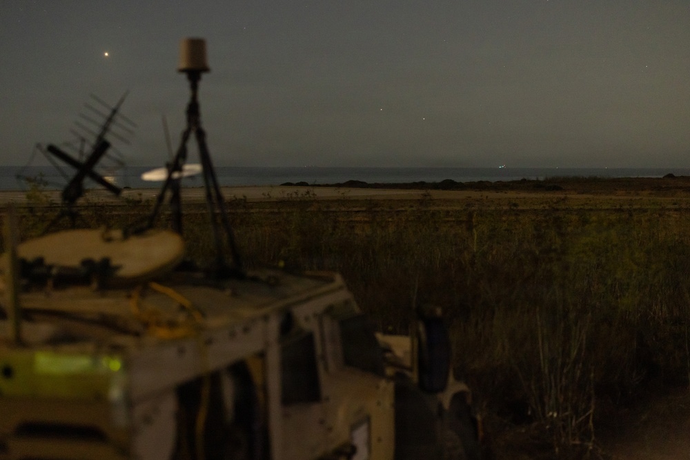 A nighttime outdoor scene showing a military or tactical vehicle in the foreground with communications equipment mounted on top. The background reveals an open flat terrain, possibly near a coastline or large body of water, with distant lights visible on the horizon. The image is taken with a long exposure, resulting in some motion blur on rotating equipment.