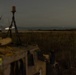 A nighttime outdoor scene showing a military or tactical vehicle in the foreground with communications equipment mounted on top. The background reveals an open flat terrain, possibly near a coastline or large body of water, with distant lights visible on the horizon. The image is taken with a long exposure, resulting in some motion blur on rotating equipment.