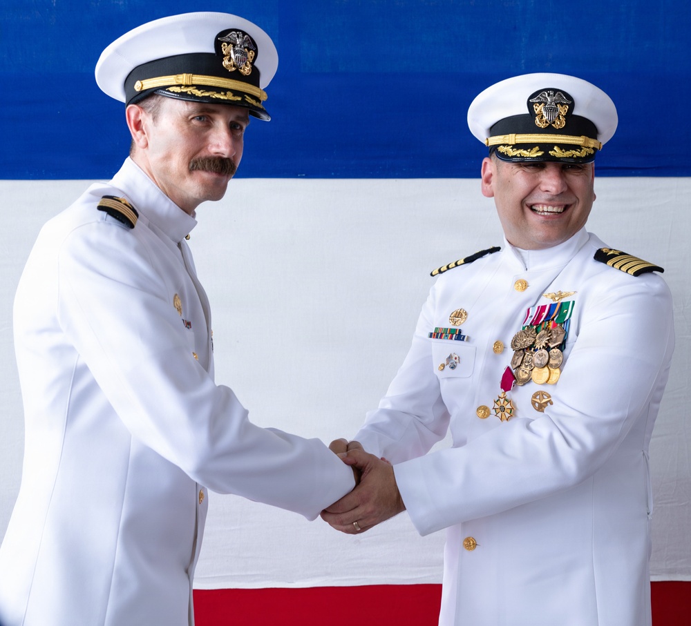 Two U.S. Navy officers in white dress uniforms are shaking hands in what appears to be a formal military ceremony, likely a change of command or retirement event. The officer on the right displays numerous medals and decorations including what appears to be a Legion of Merit medal. A red, white, and blue backdrop is visible behind them.