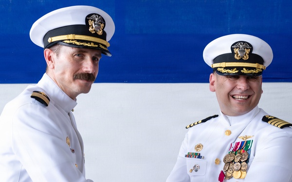 Two U.S. Navy officers in white dress uniforms are shaking hands in what appears to be a formal military ceremony, likely a change of command or retirement event. The officer on the right displays numerous medals and decorations including what appears to be a Legion of Merit medal. A red, white, and blue backdrop is visible behind them.