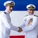Two U.S. Navy officers in white dress uniforms are shaking hands in what appears to be a formal military ceremony, likely a change of command or retirement event. The officer on the right displays numerous medals and decorations including what appears to be a Legion of Merit medal. A red, white, and blue backdrop is visible behind them.