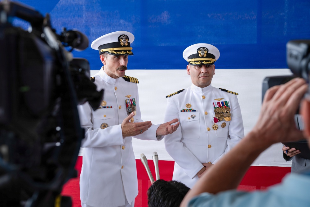 Two U.S. Navy officers in white dress uniforms stand before a red, white, and blue backdrop during what appears to be a formal military ceremony. The officer on the left is speaking with hand gestures while both display multiple medals and ribbons on their uniforms. A television camera and audience members are visible in the foreground, indicating a media-covered official event.