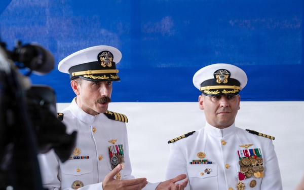 Two U.S. Navy officers in white dress uniforms stand before a red, white, and blue backdrop during what appears to be a formal military ceremony. The officer on the left is speaking with hand gestures while both display multiple medals and ribbons on their uniforms. A television camera and audience members are visible in the foreground, indicating a media-covered official event.