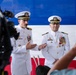 Two U.S. Navy officers in white dress uniforms stand before a red, white, and blue backdrop during what appears to be a formal military ceremony. The officer on the left is speaking with hand gestures while both display multiple medals and ribbons on their uniforms. A television camera and audience members are visible in the foreground, indicating a media-covered official event.