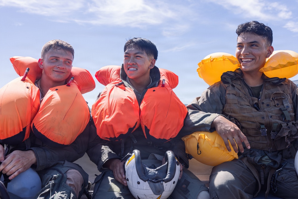 Three young military personnel are seated together, wearing inflated life vests and flight suits, appearing wet as if recently recovered from water. They are smiling and appear to be in good spirits following what appears to be a water survival training exercise. Two of the individuals are holding flight helmets, reinforcing the aviation context.