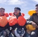 Three young military personnel are seated together, wearing inflated life vests and flight suits, appearing wet as if recently recovered from water. They are smiling and appear to be in good spirits following what appears to be a water survival training exercise. Two of the individuals are holding flight helmets, reinforcing the aviation context.