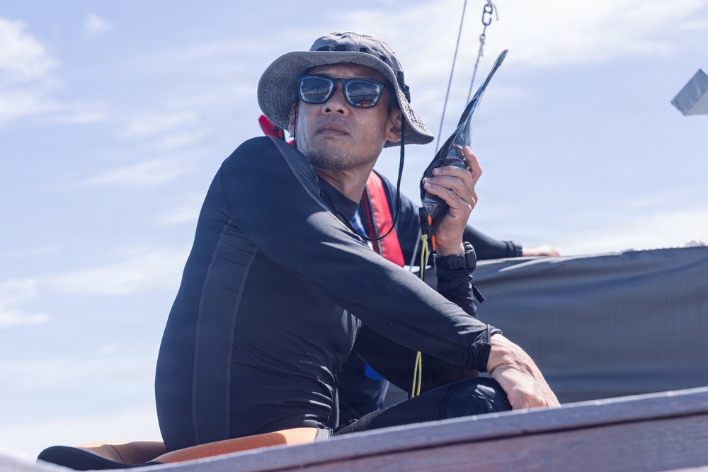 A man is seated on what appears to be a boat, holding a handheld radio or walkie-talkie near his face. He is dressed in a black wetsuit or rash guard, wearing a wide-brimmed bucket hat, sunglasses, and a red inflatable life vest. The image is taken from a low angle against a partly cloudy sky, suggesting an outdoor maritime setting.