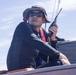 A man is seated on what appears to be a boat, holding a handheld radio or walkie-talkie near his face. He is dressed in a black wetsuit or rash guard, wearing a wide-brimmed bucket hat, sunglasses, and a red inflatable life vest. The image is taken from a low angle against a partly cloudy sky, suggesting an outdoor maritime setting.