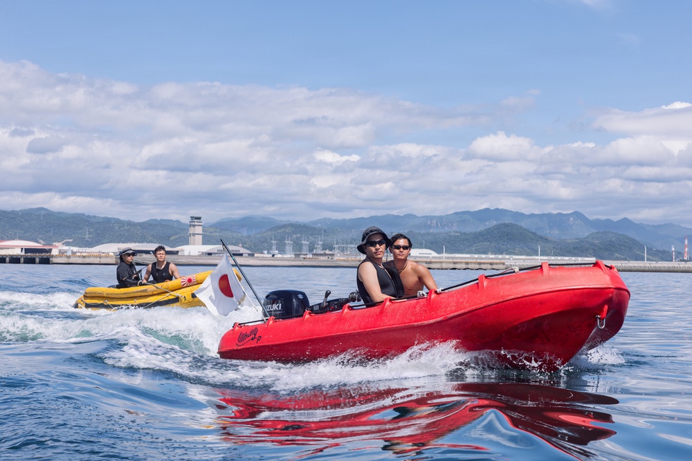 Two small motorboats are speeding across calm blue water, with mountains and industrial port infrastructure visible in the background. The foreground features a red rigid inflatable boat (RIB) with a Suzuki outboard motor carrying two passengers, while a yellow inflatable boat follows behind carrying two others. A Japanese flag (Hinomaru) is mounted on a pole between the two vessels.