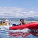 Two small motorboats are speeding across calm blue water, with mountains and industrial port infrastructure visible in the background. The foreground features a red rigid inflatable boat (RIB) with a Suzuki outboard motor carrying two passengers, while a yellow inflatable boat follows behind carrying two others. A Japanese flag (Hinomaru) is mounted on a pole between the two vessels.